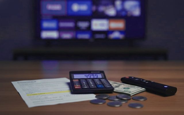 A calculator, dollar bills, coins, a streaming stick and a remote on a table in front of a TV